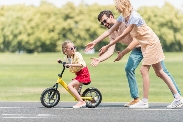 happy parents teaching little daughter riding bicycle in park - Foto, imagen