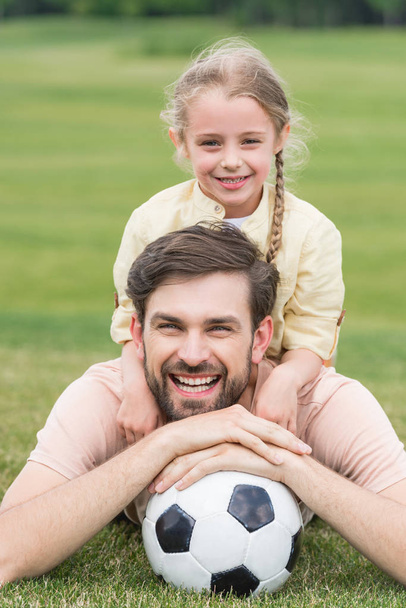 glücklicher Vater und Tochter lächeln in die Kamera, während sie mit Fußballball auf dem Rasen liegen - Foto, Bild