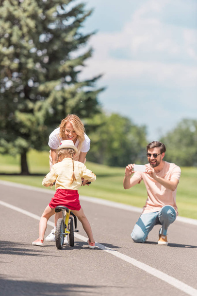 happy young man with smartphone photographing smiling mother teaching daughter riding bicycle in park - Foto, imagen