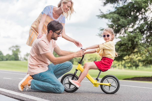 счастливые родители смотрят на маленькую дочь на велосипеде в парке
  - Фото, изображение