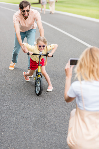 woman with smartphone photographing happy father and daughter riding bicycle in park  - Fotoğraf, Görsel