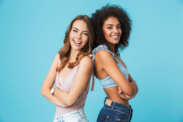 Two smiling young girls dressed in summer clothes standing back to back and looking at camera isolated over blue background - Foto, immagini