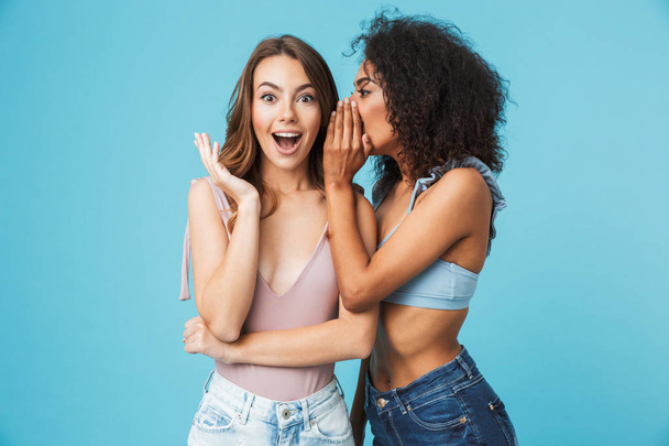 Two cheerful young girls dressed in summer clothes whispering secrets isolated over blue background - Foto, Bild