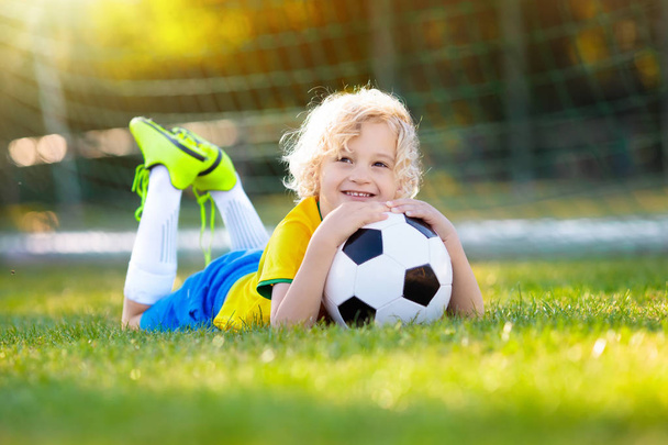Kids play football on outdoor field. Brazil team fans. Children score a goal at soccer game. Little boy in Brazilian jersey and cleats kicking ball. Football pitch. Sports training for player. - Zdjęcie, obraz