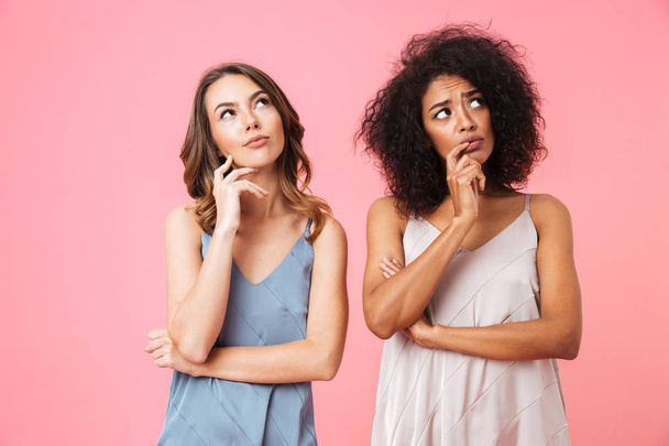 Two pensive young girls dressed in summer clothes looking away at copy space isolated over pink background - Foto, imagen