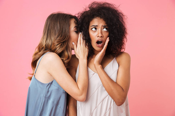 Two shocked young girls dressed in summer clothes whispering secrets isolated over pink background - Foto, immagini
