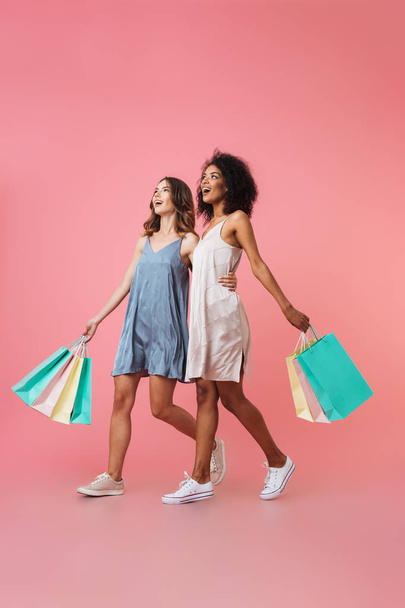Full length portrait of two smiling young girls dressed in summer clothes holding shopping bags while walking isolated over pink background - Φωτογραφία, εικόνα