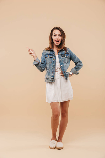 Full length portrait of an excited young girl in summer clothes pointing at copy space isolated over beige background - 写真・画像