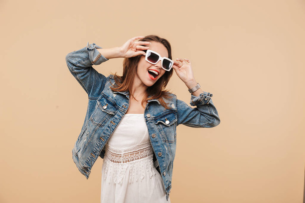 Portrait of a happy young girl in summer clothes and sunglasses posing while standing isolated over beige background - Фото, изображение