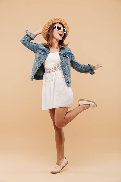 Full length portrait of a cheerful young girl in summer clothes and sunglasses posing while standing isolated over beige background - Photo, Image