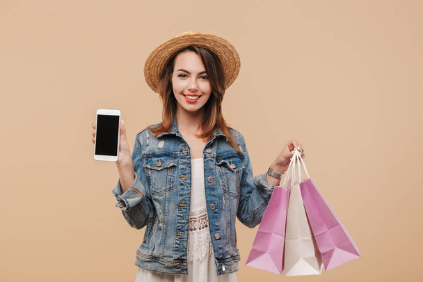 Portrait of a cheerful young girl in summer clothes showing blank screen mobile phone while holding shopping bags isolated over beige background - Zdjęcie, obraz