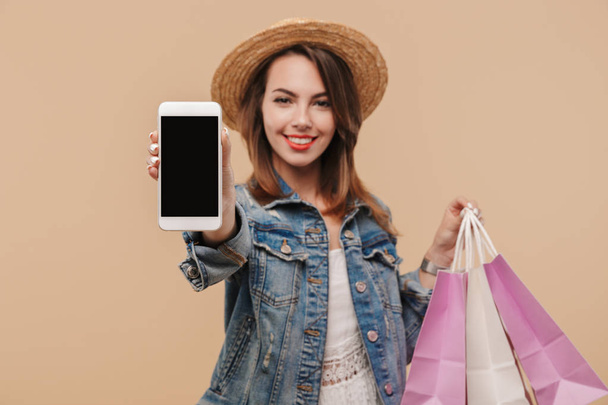 Portrait of a happy young girl in summer clothes showing blank screen mobile phone while holding shopping bags isolated over beige background - Фото, изображение