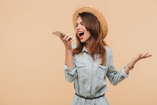 Portrait of a furious young girl in summer clothes yelling at mobile phone isolated over beige background - Фото, изображение