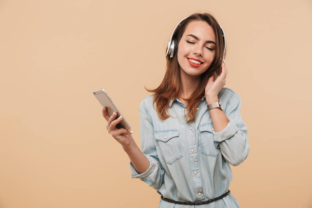 Portrait of a smiling young girl in summer clothes listening to music with headphones while holding mobile phone isolated over beige background - Foto, Bild