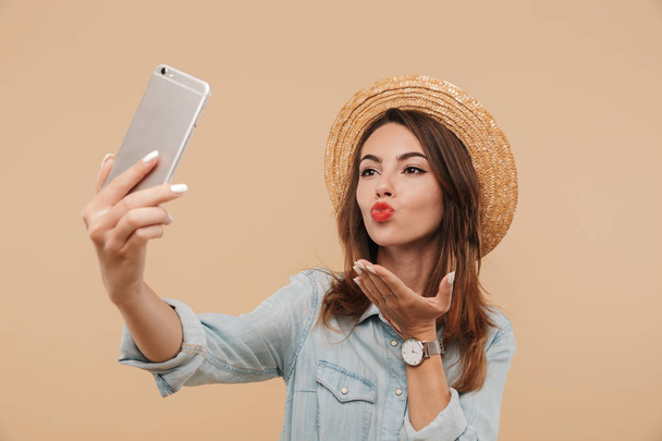 Portrait of a pretty young girl in summer clothes taking a selfie and sending kiss isolated over beige background - Фото, изображение