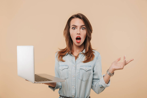 Portrait of a surprised young girl in summer clothes holding laptop computer isolated over beige background - Foto, afbeelding