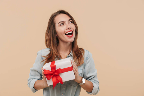 Portrait of a happy young girl holding present box and looking at copy space isolated over beige background - Фото, изображение