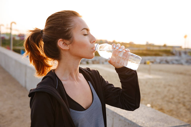 Image of attractive beautiful young sports woman drinking water outdoors on the street. - 写真・画像