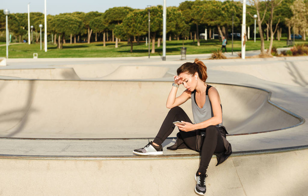 Image of young tired sports lady sitting outdoors in park using mobile phone looking aside listening music. - Photo, image