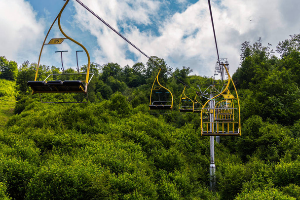 Seilbahn mit Kabinen für drei Sitze und gelbem Geländer vor dem Hintergrund saftig grüner Bäume - Foto, Bild