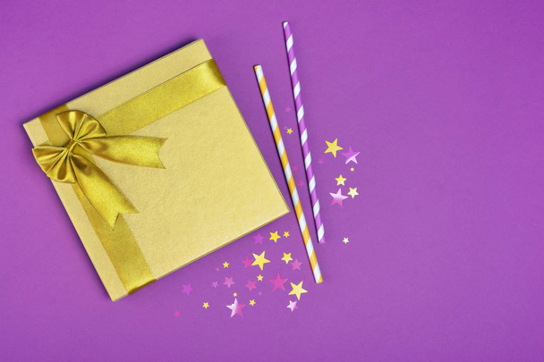 Golden shiny classic gift box with satin bow and cocktail straws with confetti in the shape of stars as attributes of party on purple background - Фото, зображення