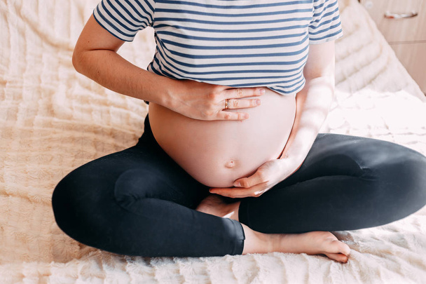 T シャツで妊娠中の女性は、ウィンドウに対して自宅で白い背景の上の腹に手を保持します。妊娠、出産準備、期待のコンセプトです。クローズ アップ, コピー スペース, 屋内で。妊娠の美しい柔らかい雰囲気写真. - 写真・画像