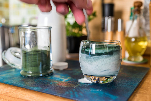 Nahaufnahme Ansicht frisches Kefir Probiotik Getränk in klarem Glas und Löffel mit grünem Spirulinapulver bereit, auf dem Küchentisch zu mischen - Foto, Bild