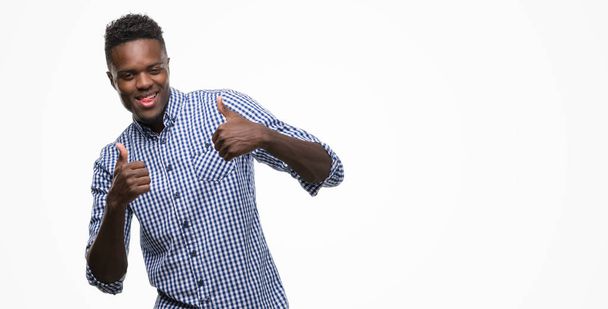 Młody człowiek african american na sobie niebieską koszulę znak sukcesu robi pozytywny gest ręką, kciuki, uśmiechnięty i zadowolony. Patrząc na kamery z wesoły wyrażenie, zwycięzca gest. - Zdjęcie, obraz