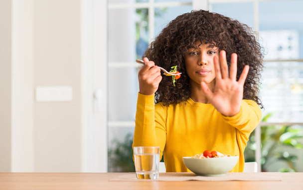 Afrikkalainen amerikkalainen nainen syö pastasalaattia kotona avoimella kädellä tekemällä stop-merkin vakavalla ja itsevarmalla ilmeellä, puolustus ele
 - Valokuva, kuva