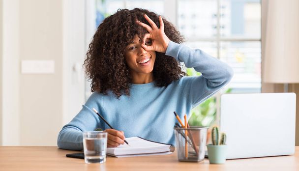 African american woman studying at home using a computer laptop with happy face smiling doing ok sign with hand on eye looking through fingers - 写真・画像