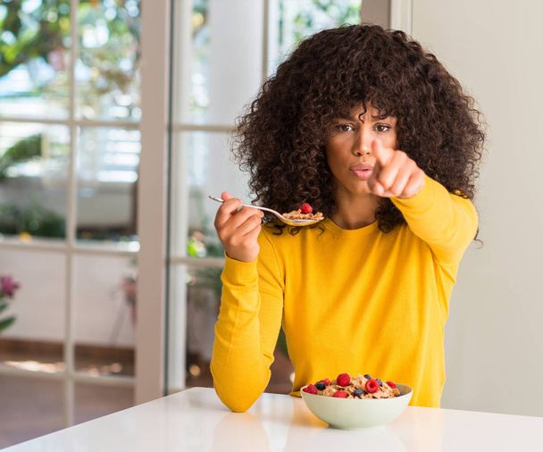 Mujer afroamericana comiendo cereales, frambuesas y arándanos señalando con el dedo a la cámara y a ti, signo de mano, gesto positivo y seguro desde el frente
 - Foto, Imagen