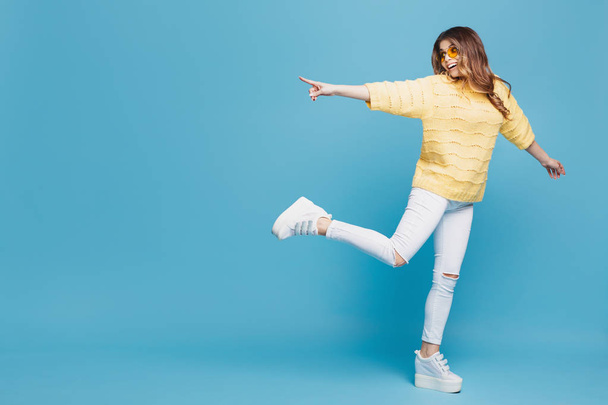 Pretty young woman in yellow sweater posing on blue background. Attractive woman pointing fingers to the left with cheerful and happy expression. Copy space. - 写真・画像