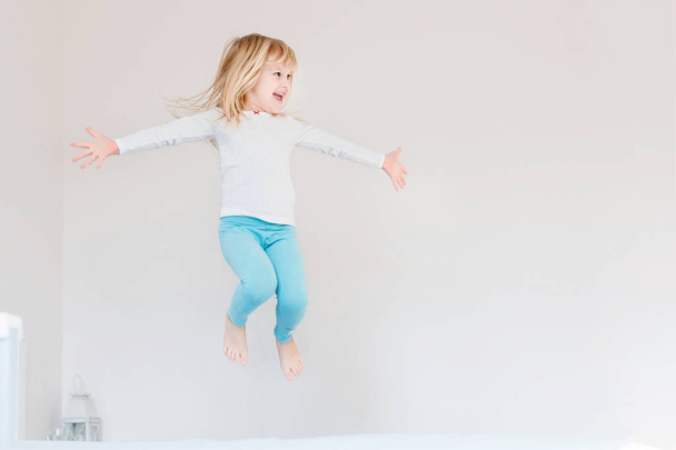 Happy kid jumping over bed. Cute little blond girl having fun indoors. Happy and careless childhood concept - Foto, Imagem