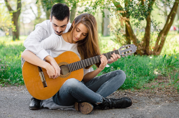 Νεαρός άνδρας δείχνει όμορφο θηλυκό φίλο του πώς να παίξει ακουστική κιθάρα στο πάρκο - Φωτογραφία, εικόνα