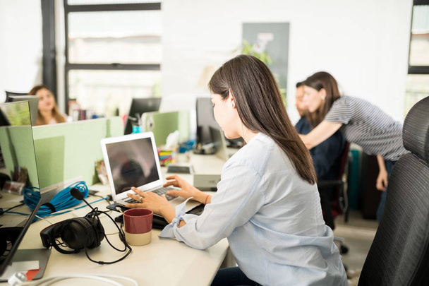Giovane donna d'affari che lavora al computer alla scrivania dell'ufficio con colleghi sullo sfondo
  - Foto, immagini