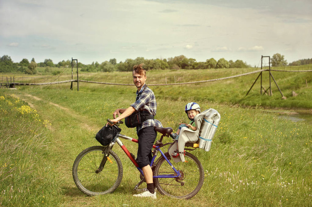 Kırsal Bisiklet seyahat. Baba bisiklet ve bebek sandalye seyahat suspenced Köprüsü yakınlarında kırsal yolda yürümeye başlayan çocuk oğlu. Etkin Aile kavramı. - Fotoğraf, Görsel