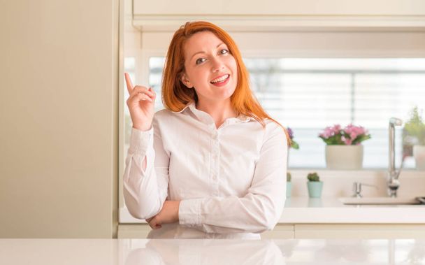 Roodharige vrouw in de keuken met een grote glimlach op het gezicht, met de hand en de vinger te wijzen naar de kant te kijken naar de camera. - Foto, afbeelding