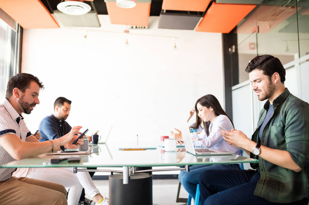 Grupo de empresarios sentados alrededor de la mesa de conferencias y utilizando sus teléfonos móviles mientras ignoran el trabajo y las redes sociales
. - Foto, imagen