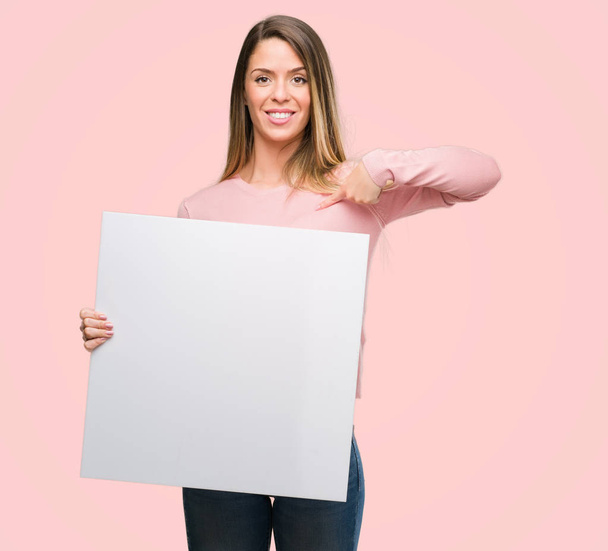 Beautiful young woman holding advertising banner with surprise face pointing finger to himself - Foto, Bild