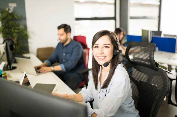 Retrato de una mujer latina bastante joven con auriculares sentados en el escritorio de la oficina con compañeros de trabajo en el fondo
 - Foto, imagen