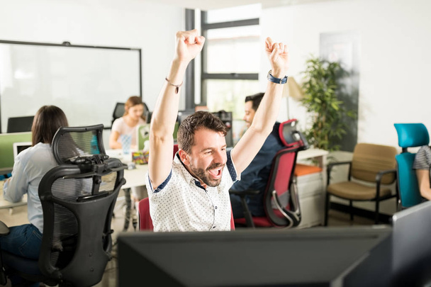 Портрет веселого позитивного чоловіка в каузальному вбранні, що піднімає руки і святкує досягнення, сидячи в сучасному офісі з колегами на фоні
  - Фото, зображення
