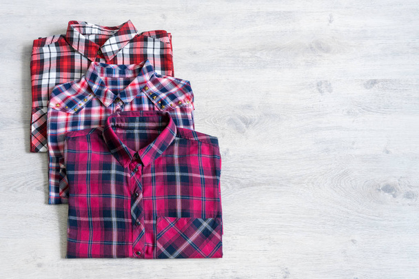 Three multi-colored checkered women's shirts lying neatly folded on a light wooden background - 写真・画像