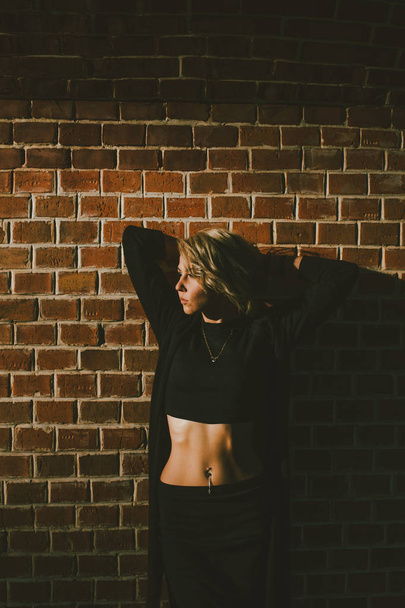 Красивая блондинка с пирсингом на пупке, в топике, стоящем у кирпичной стены, руки за головой, на закате
 - Фото, изображение