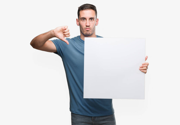 Handsome young man holding advertising banner with angry face, negative sign showing dislike with thumbs down, rejection concept - Fotografie, Obrázek