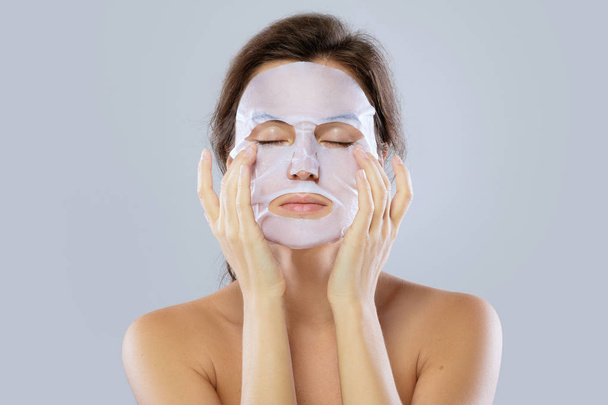 Face care and beauty treatments. Woman with a cloth moisturizing mask on her face isolated on gray background - Fotó, kép