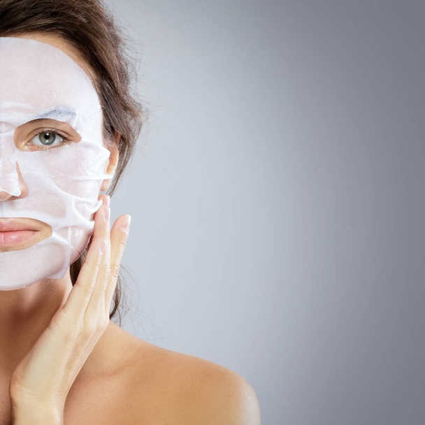 Face care and beauty treatments. Woman with a cloth moisturizing mask on her face isolated on gray background - Фото, изображение