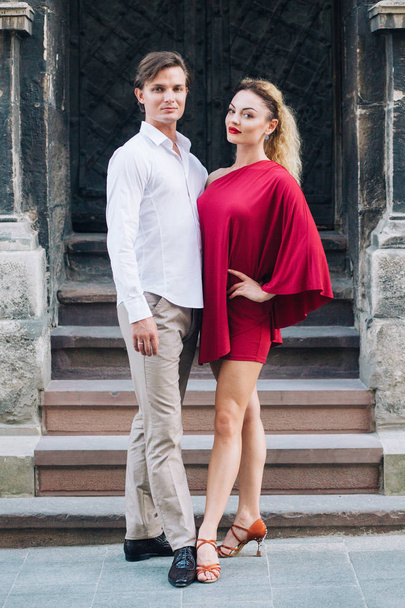 beautiful happy couple in love posing at old building in sunny street. stylish hipster groom and blonde bride in red dress gently hugging, professional dancers. romantic moments in summer - Fotó, kép