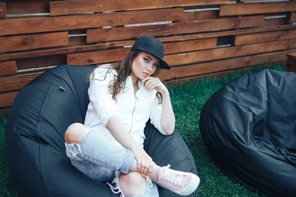 Young woman in a black cap sits on a black puff outdoor. Black bean bag chair outdoor - Valokuva, kuva