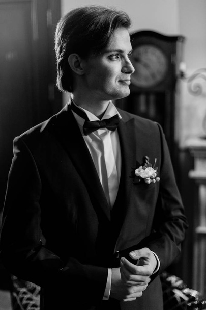 stylish groom in bow tie and blue suit getting ready in the morning in hotel, looking in mirror. wedding day concept. handsome man preparations in vintage room, in tuxedo with boutonniere - Photo, Image