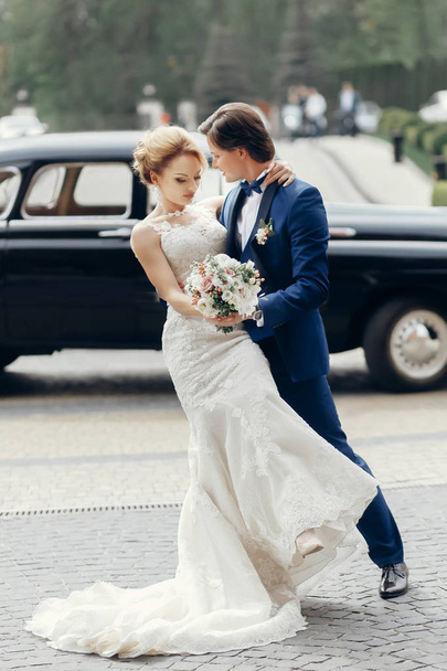 Luxus-Hochzeitspaar tanzt an altem Auto im Licht. Stilvolles Brautpaar, das sich auf der Straße umarmt und umarmt. romantische leidenschaftliche sinnliche Moment. Frau und Mann zusammen - Foto, Bild